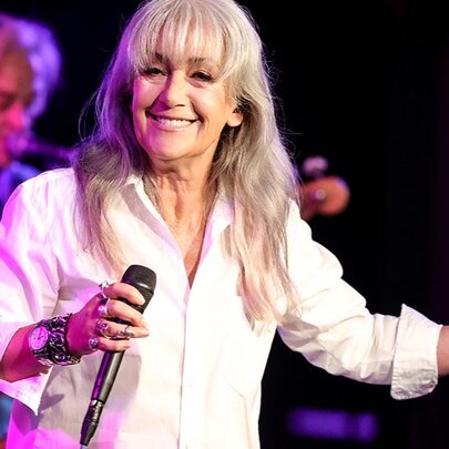 Singer Wendy Matthews smiling warmly on stage holding a microphone. 