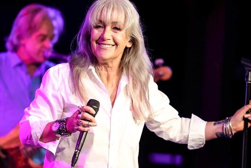Singer Wendy Matthews smiling warmly on stage holding a microphone. 