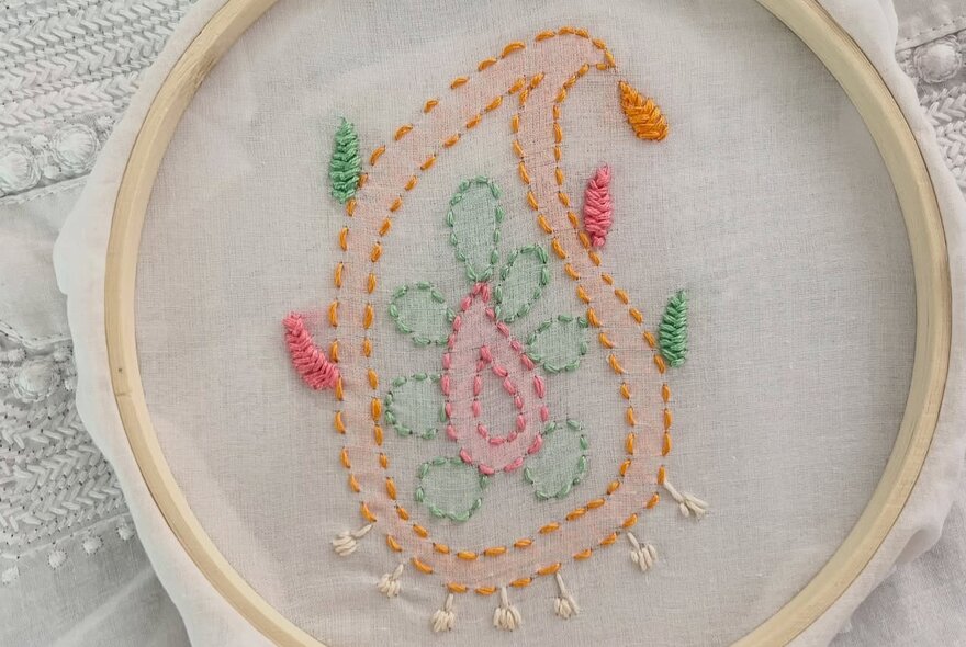A white piece of cotton with pale coloured embroidered stitching, held taut in a circular wooden frame.