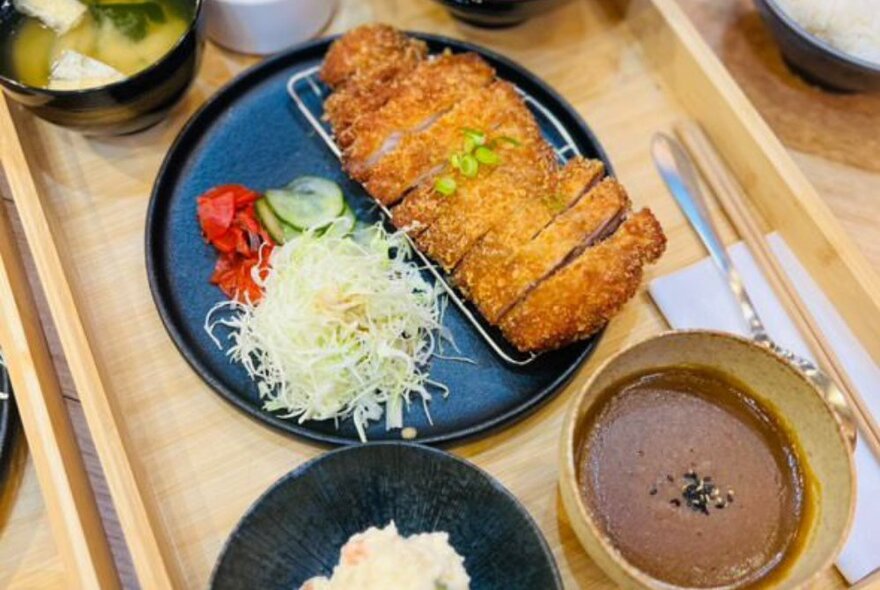 A plate of fried sliced pork Japanese style, pickled daikon, sauces and rice, on a wooden tray on a restaurant table.