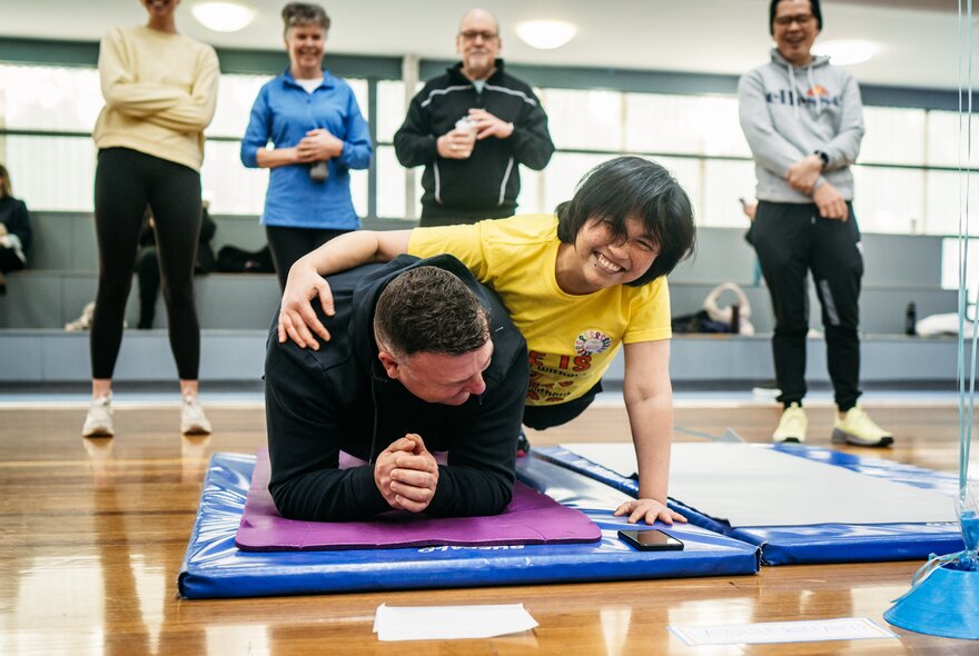 Two people doing planks on exercise mats in an indoor hall, surrounded by standing spectators.