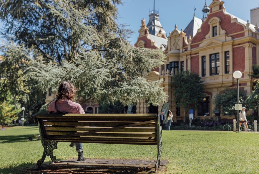 A person seated by themselves on an outdoor seat, on a lawn, with a stately building in the background.