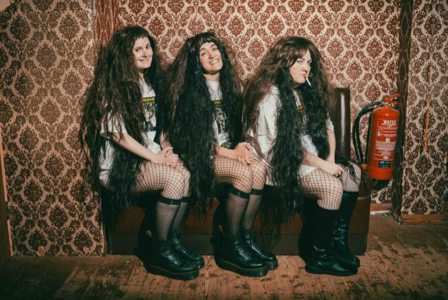 A scene from the theatre show Philosophy of the World with three people posed for a photo on a bench against patterned wallpaper - they wear long black hair wigs, oversized white t-shirts, black knee-high socks and boots.