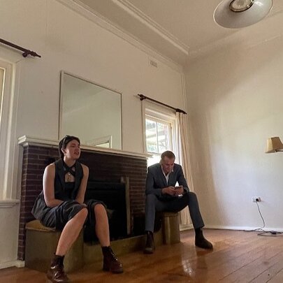 People seated in an empty room with a fireplace and lamp.