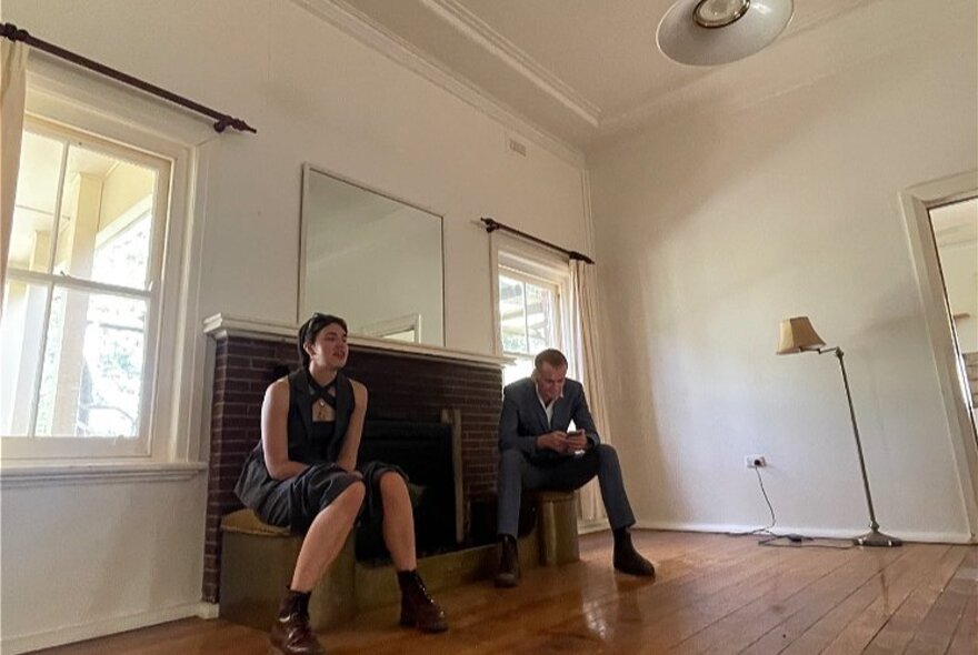 People seated in an empty room with a fireplace and lamp.