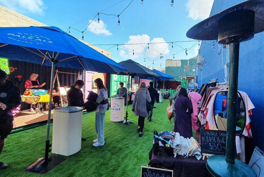 Outdoor courtyard space at EziStreat food hall, with large blue market umbrellas, green astroturf and people walking around.