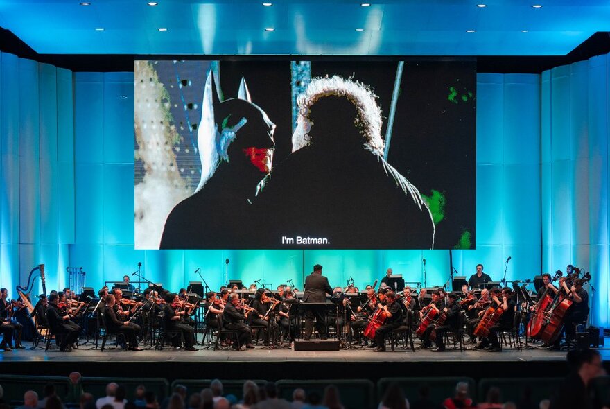 Huge screen showing Batman movie suspended over massed choir on stage.