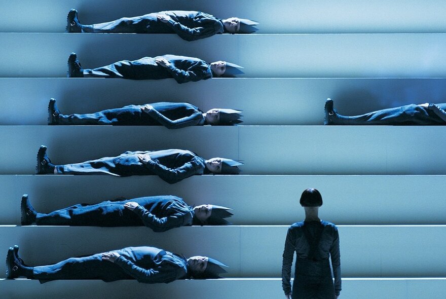 Abstract image in blue tones of a person looking at a staircase structure with a person laying on each stair. 