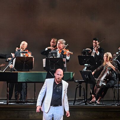 A singer wearing a light coloured suit standing in front of a performing orchestra on stage and singing.