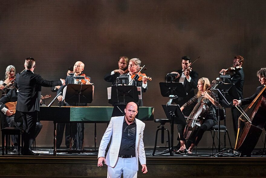 A singer wearing a light coloured suit standing in front of a performing orchestra on stage and singing.