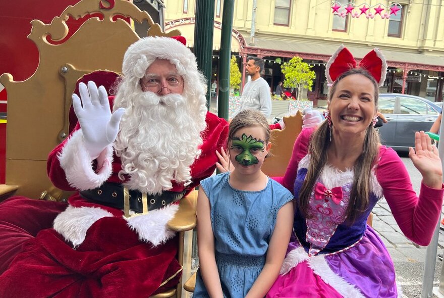 A girl with face paint and a young woman in a princess outfit pose with Santa.