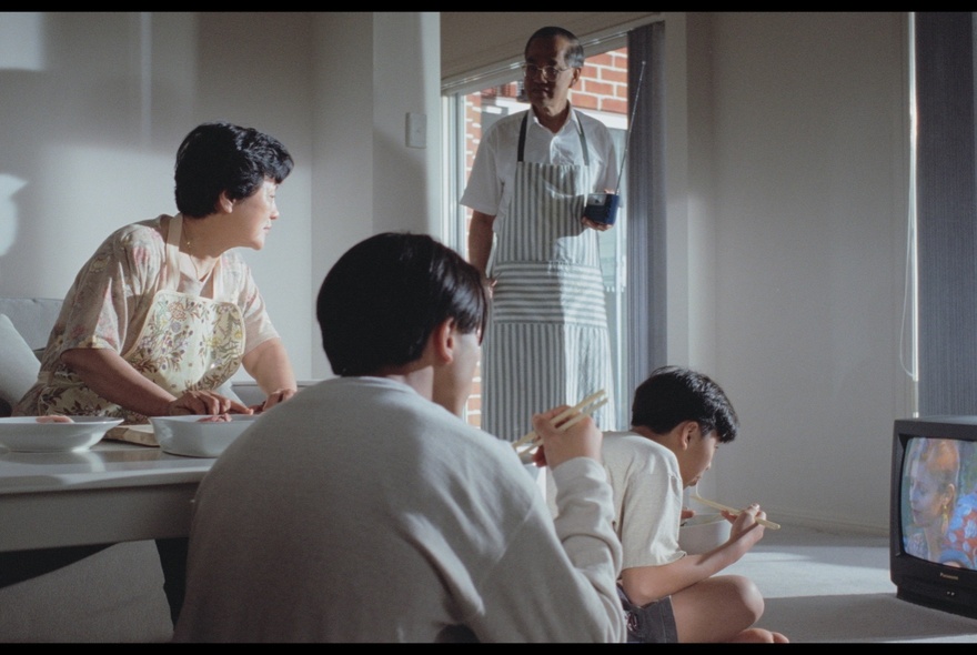 A still from the film Floating Life showing a family interacting with a dad coming through the door wearing an apron and kids watching tv while eating. 
