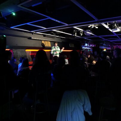 Performer on a stage in front of an audience in a dark venue.