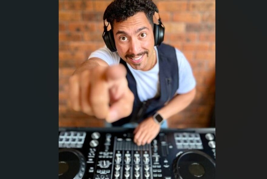A DJ wearing headphones behind a mixing desk, smiling and pointing his finger at the camera so it's blurred and foreshortened.