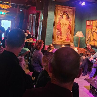 A person holding a microphone performing stand-up comedy on a small stage at Fad Gallery Bar, in front of a live audience.