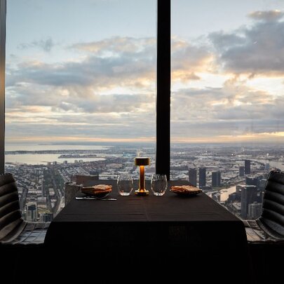 Interior view of Eureka 89 with a dining table and two chairs in the foreground positioned against a large window, with a panorama of Melbourne city and the bay visible in the background.