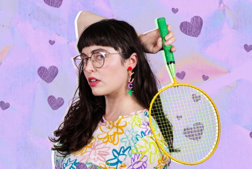 A person with long brown hair, wearing glasses and a top with a floral design, holding a small racquet behind her head, posed against a purple background.