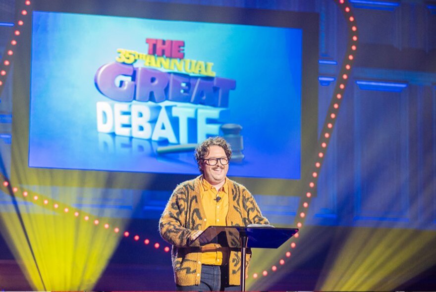 A smiling comedian standing at a lectern on a stage, behind them the words THE GREAT DEBATE are projected onto a screen.