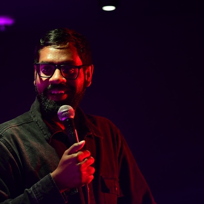 A middle aged man talking into a microphone he holds in his hand, performing stand-up comedy on a darkened stage.