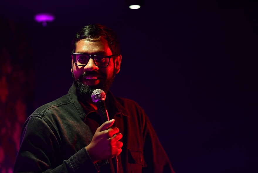 A middle aged man talking into a microphone he holds in his hand, performing stand-up comedy on a darkened stage.