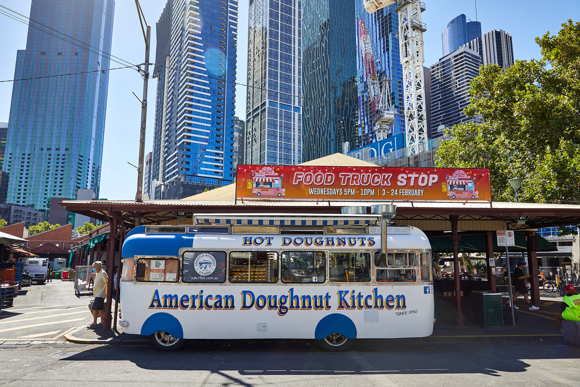A van parked at a city market with the name American Doughnut Kitchen painted on the side.