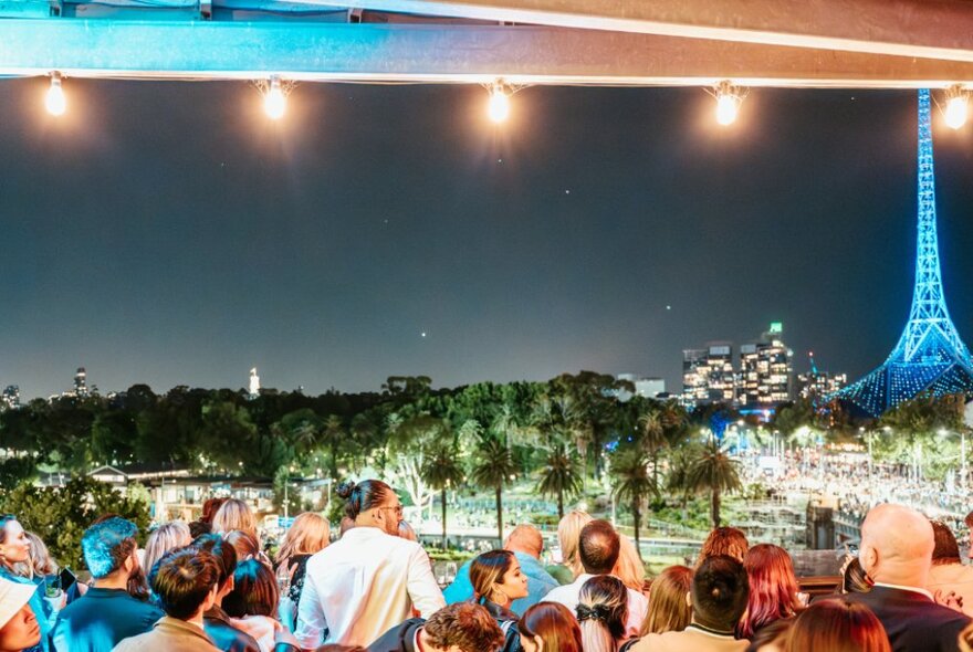 People on Transit Rooftop Bar looking across to the blue illuminated Arts Centre at night. 