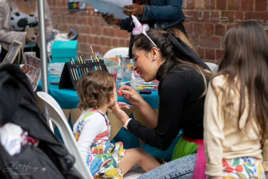 A young child having her face painted by a woman wearing a unicorn-horn headband; another child waiting patiently for her turn.