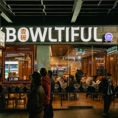 Outside a busy noodle restaurant there is a large sign above the entrance saying bowltiful