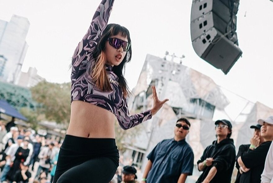 A woman wearing a psychedelic crop top and sunglasses, dancing in front of a crowd at Melbourne's Fed Square.