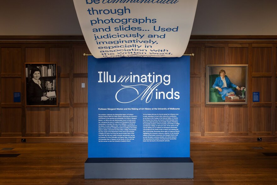 A gallery-like space with wooden wainscoting and a large blue poster standing in the middle of two hanging portraits.