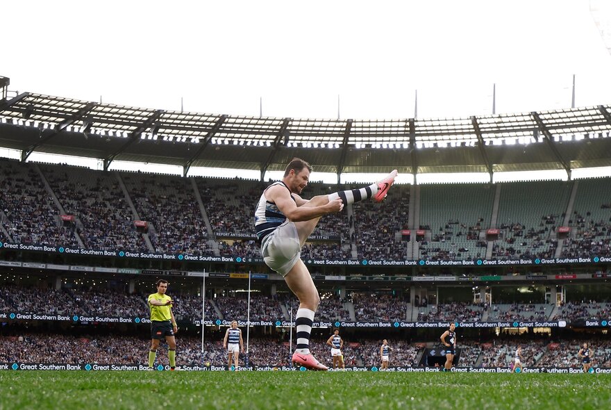 Geelong AFL football player kicking the ball during a match, with the stadium filled with spectators behind him.