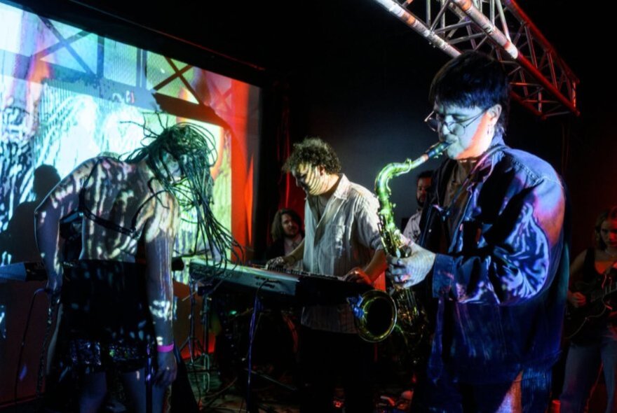 Three musicians performing on a small stage with coloured lights. 