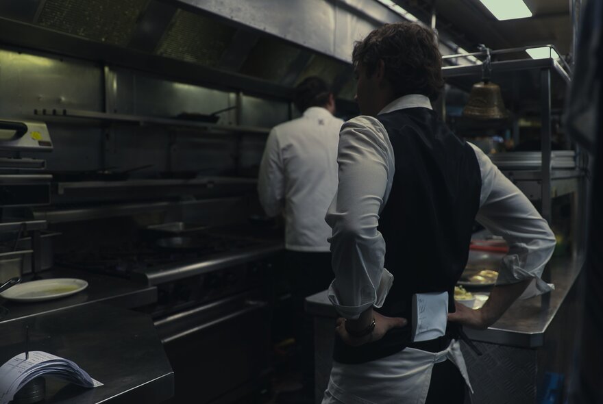A waiter standing with their hands on their hips in a commercial kitchen, with a chef in the background preparing food.