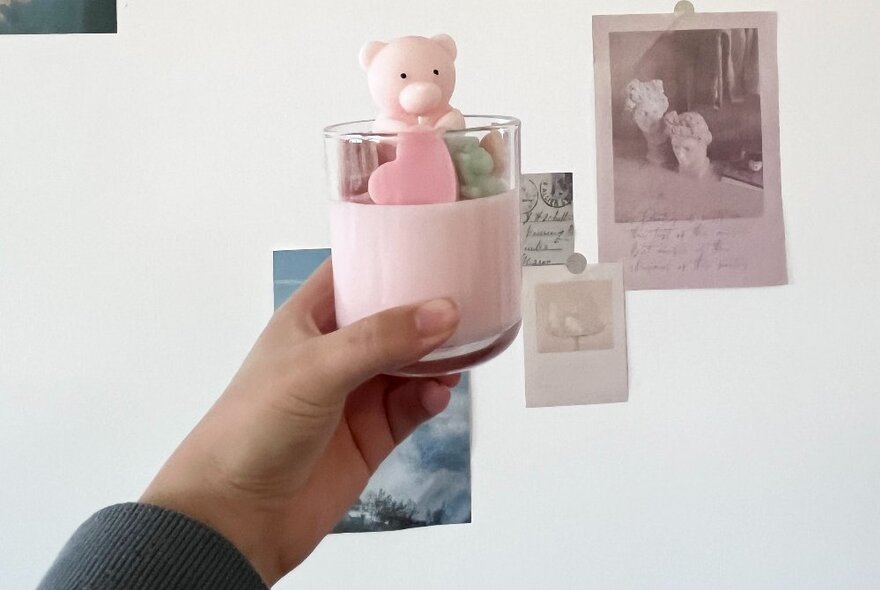 A hand holding up a pink candle in a glass with a small bear toy poking out of the top. 