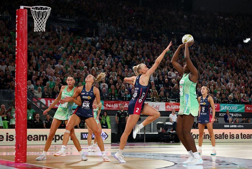 A professional netball match, with players on the court, one about to shoot for goal, and fans in the stadium seats in the background. 