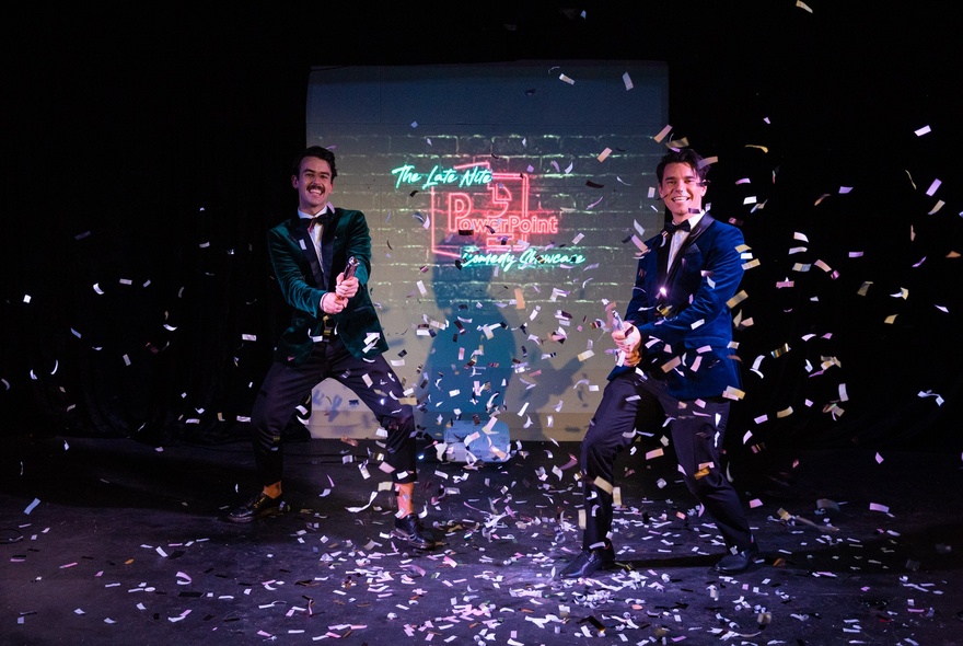 Two performers in formal jackets and bow ties stand on a stage with confetti falling around them.