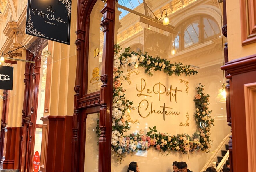 A decorated storefront inside a vintage arcade, featuring a large white floral display wall with the words 'Le Petit Chateau' written in elegant script.