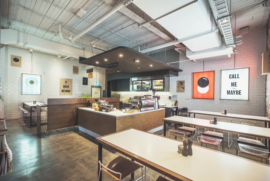 Interior of cafe showing the serving area, long communal tables and posters on a brick wall.