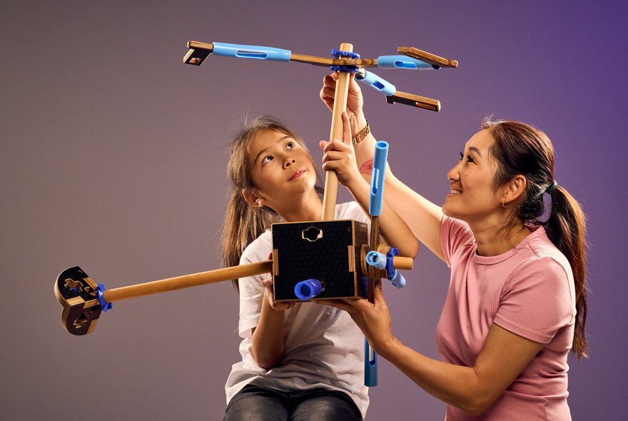 A young teenager and an adult holding up and looking at a construction they've made from pieces of timber and plastic tubing.