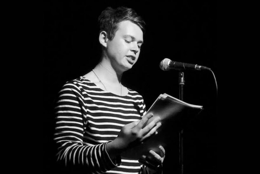 A person wearing a striped top, standing in front of a microphone and reading from a notepad they hold in their hands.