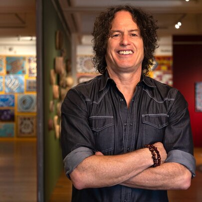 A man in a dark denim shirt standing with his arms crossed and smiling, a gallery behind him.