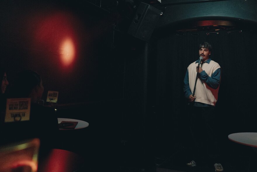 A dark image of a comedian on stage in a white jumper and black pants.