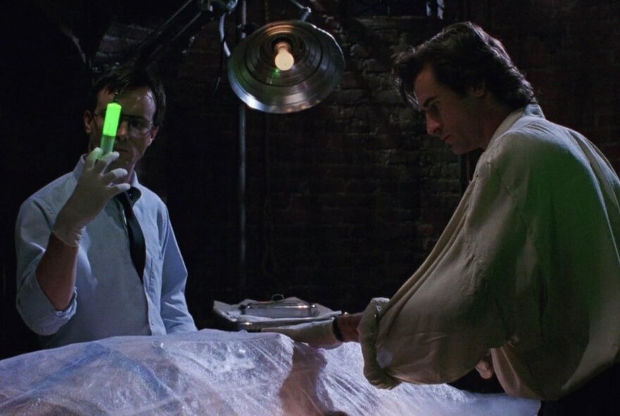 Two people with a body under a sheet, one man holding a luminous green tube.