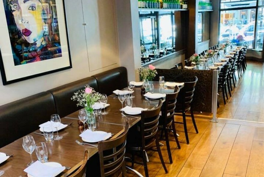 Long row of tables and chairs on wooden floor leading to front window, artwork on wall.