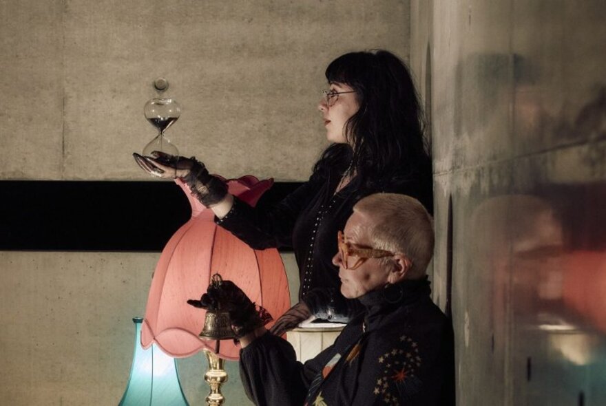 Profile of two women standing with their backs to a wall, holding out their hands with coloured lampshades in the background. 