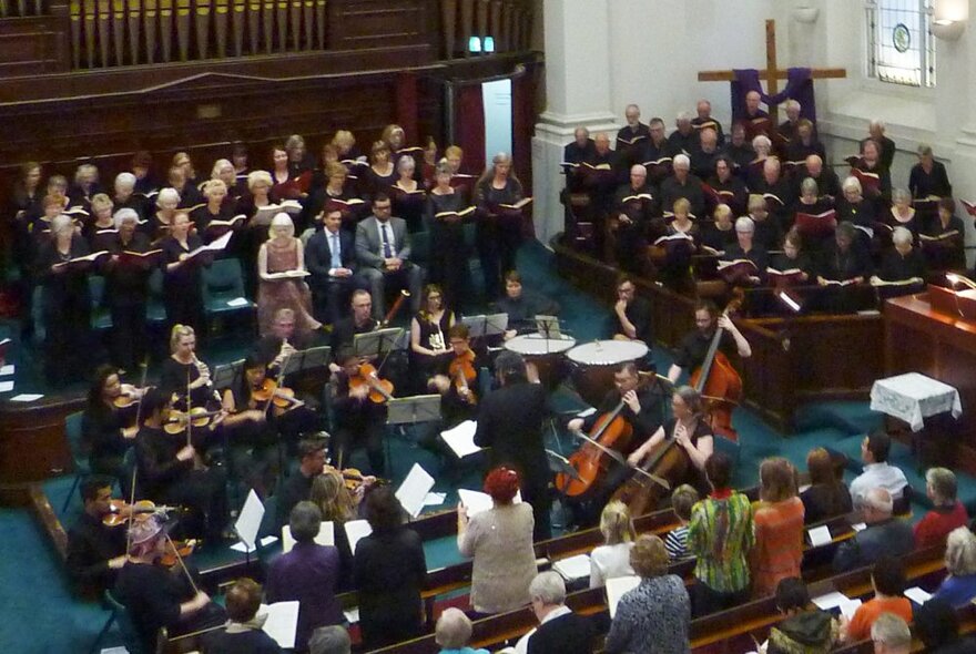 Choir and audience in a church chapel, choir standing in front of pipe organ, small orchestra with cellos and people standing in pews.