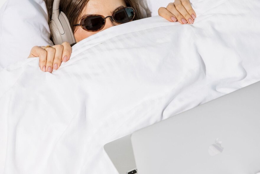 Person wearing sunglasses lying in bed with sheets pulled up to their nose, a laptop on the bed.