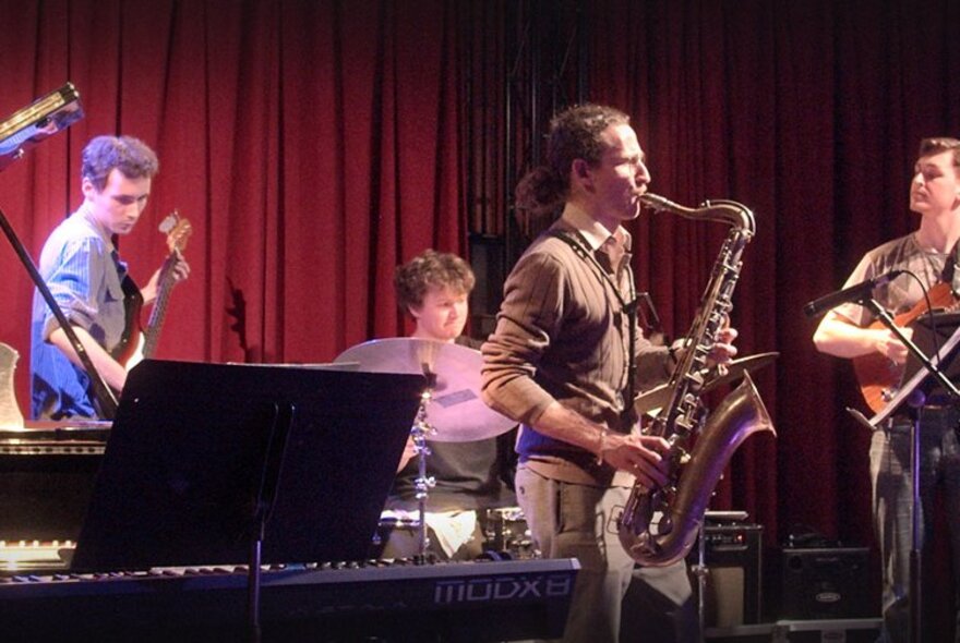 A band of musicians on a small stage all playing instruments including saxophone, drums, guitars and keyboards.