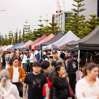 People walking past rows of outdoor market stalls with umbrellas and trees.
