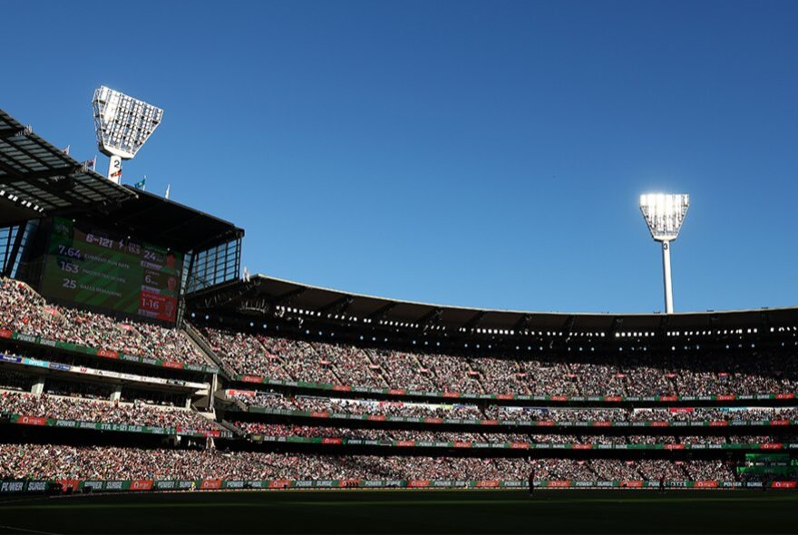 A wide-angle shot of the Melbourne Cricket Ground (MCG) grandstands filled with a large crowd under a clear, bright blue sky and two tall, stadium floodlight towers.